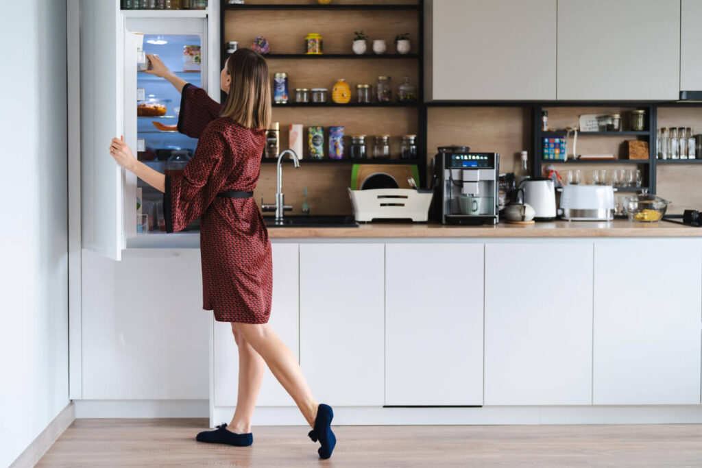 A woman in a red robe stands in a modern kitchen with smart kitchen storage, reaching into an open refrigerator. The space features white cabinets, wooden shelves with jars, and various appliances on the counter.