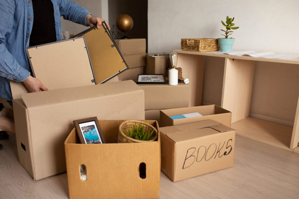 Packing for a Big Move, a person fills cardboard boxes in a room, with books, a photo frame, home decor, and a plant nearby. Some boxes are labeled, while an empty shelf stands against the wall.