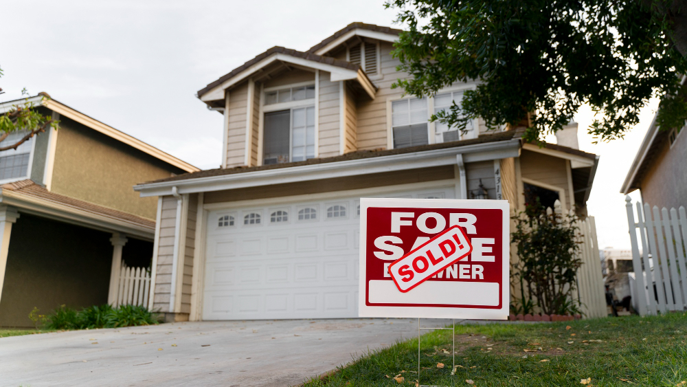 A two-story suburban house with a white garage and beige siding. A red For Sale sign in the front yard displays a SOLD! sticker, indicating the home has been purchased.