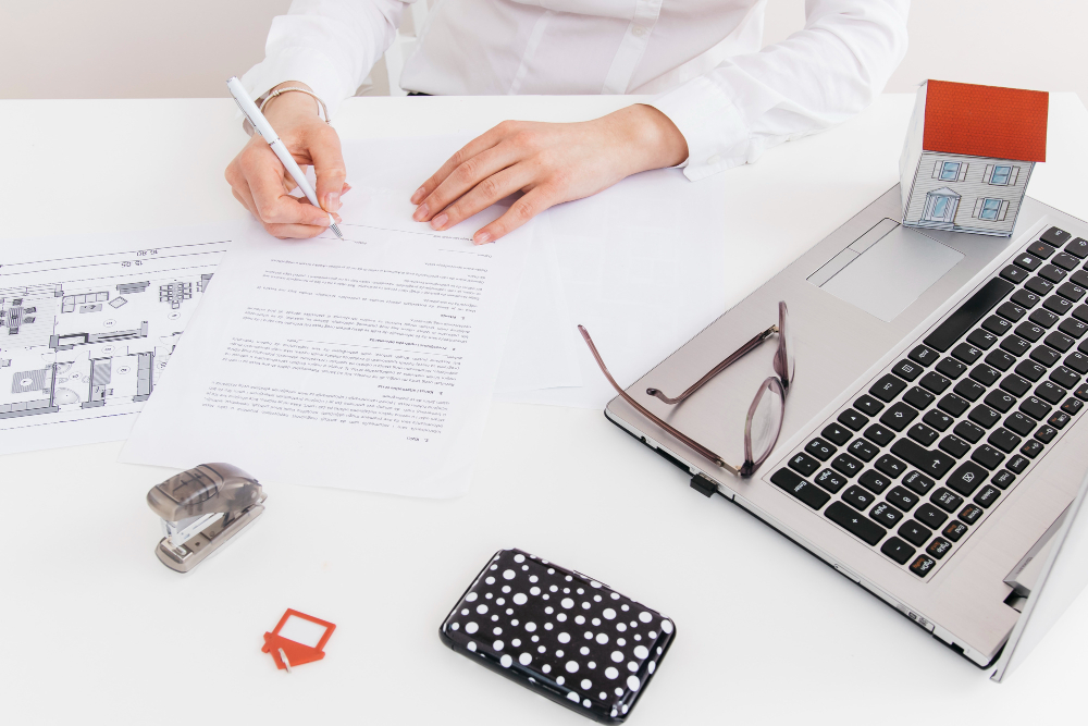 A person fills out documents at a desk with floor plans, a laptop, glasses, a small paper house, a dotted wallet, and office supplies.