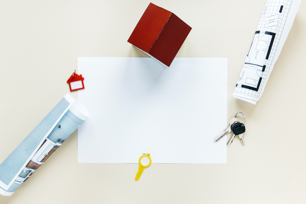 A blank sheet of paper is surrounded by a small red house model, rolled-up blueprints, a red house-shaped keychain, a yellow key, and two silver keys on a keyring, all set on a light background.
